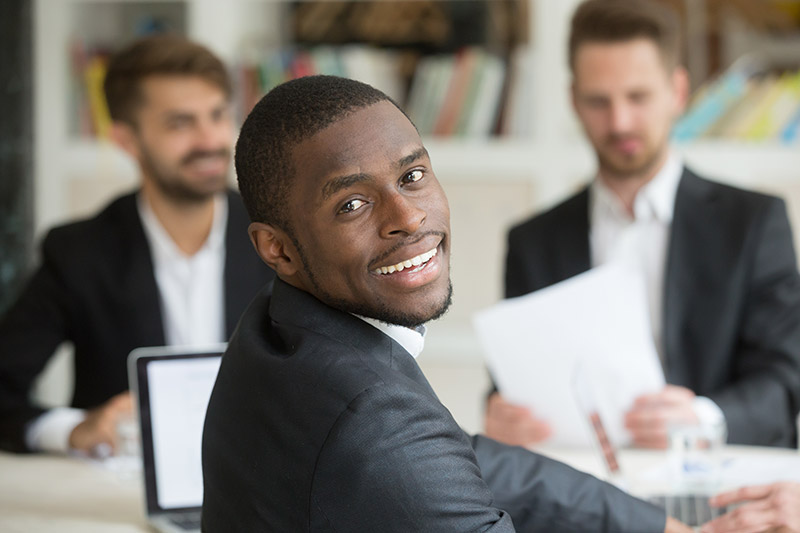 african american young male professional facing camera sitting at desk with other young professional men